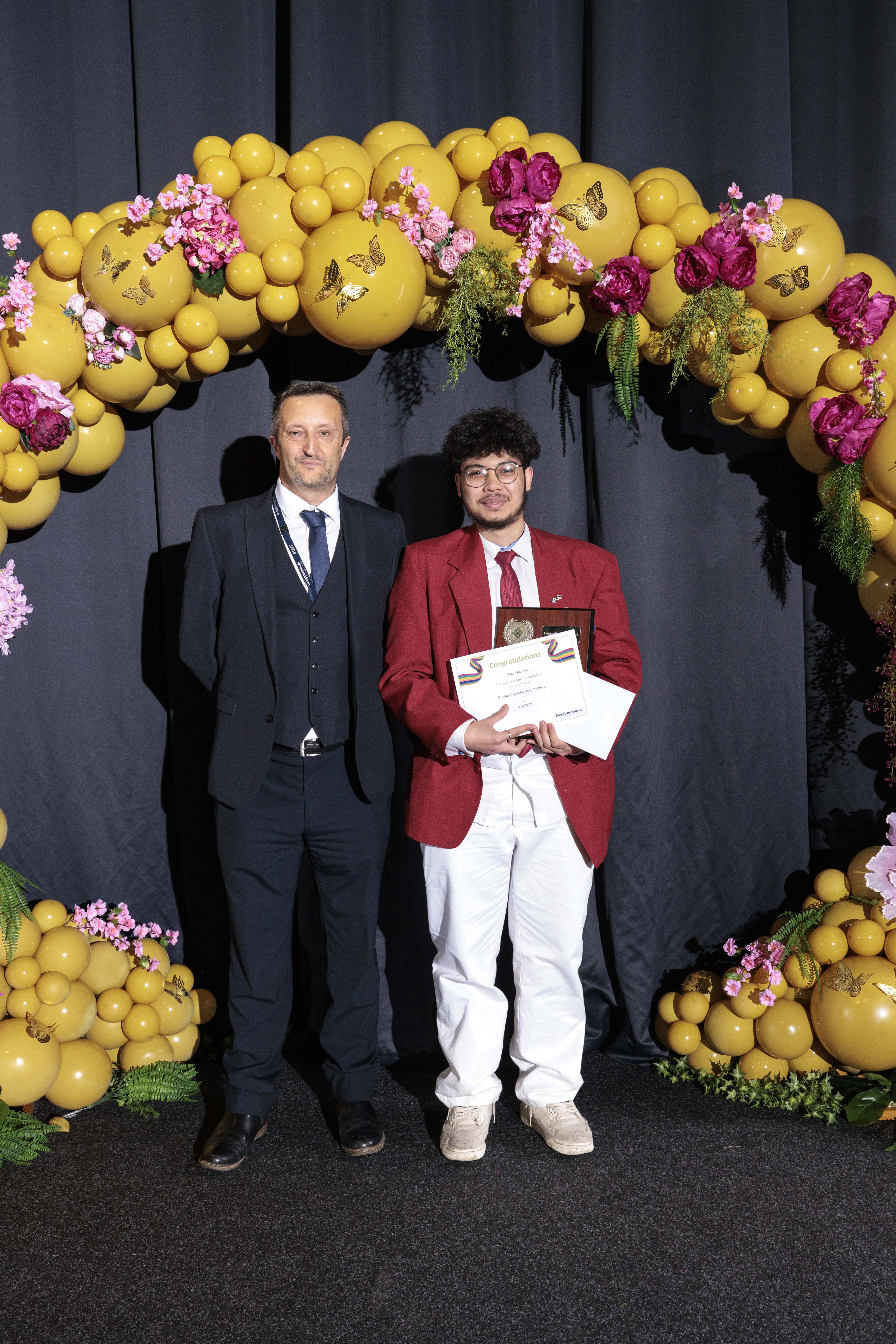 Eddie standing in front of a ballon arch in smart formal clothing, holding a certificate and award.